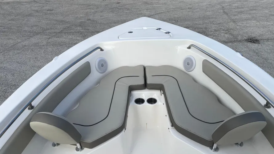 The bow seating area of a boat, featuring two cushioned seats with backrests in a V-shaped configuration. The seats are upholstered in light gray with dark gray accents, and two cup holders are visible in the center between the seats. A stainless steel grab rail lines the edges of the bow for added safety, and a hatch is visible on the deck in front of the seating area. The design provides a comfortable and spacious area for passengers to relax at the front of the boat.