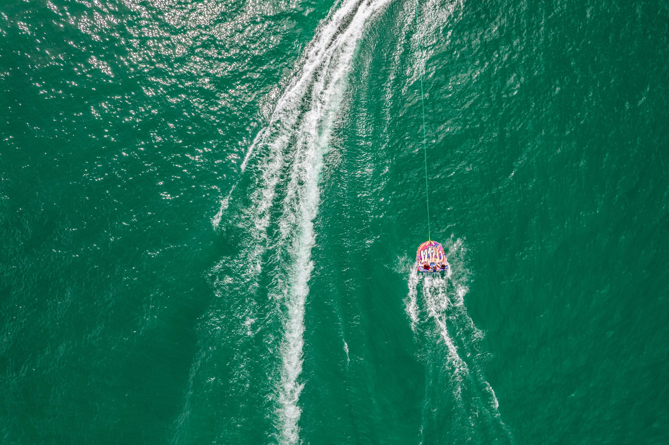 Aerial view of a group of people riding on a large inflatable tube being towed across vibrant green water. The boat towing them is out of frame, but the path of its wake is visible, creating ripples and waves in the water. The bright, multicolored inflatable tube contrasts with the water, and the riders are holding on tightly, enjoying the experience. The scene captures the excitement and fun of tubing on open water.