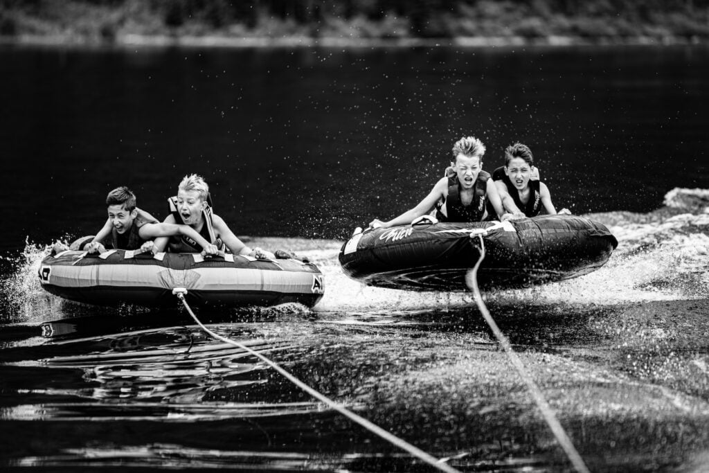 Four children ride on two towable tubes while being pulled across a lake. They are wearing life jackets and holding onto the handles of the tubes as water splashes around them. The black-and-white image captures their expressions of excitement and concentration as they enjoy the thrill of tubing. The tubes are gliding across the water's surface, leaving ripples in their wake.