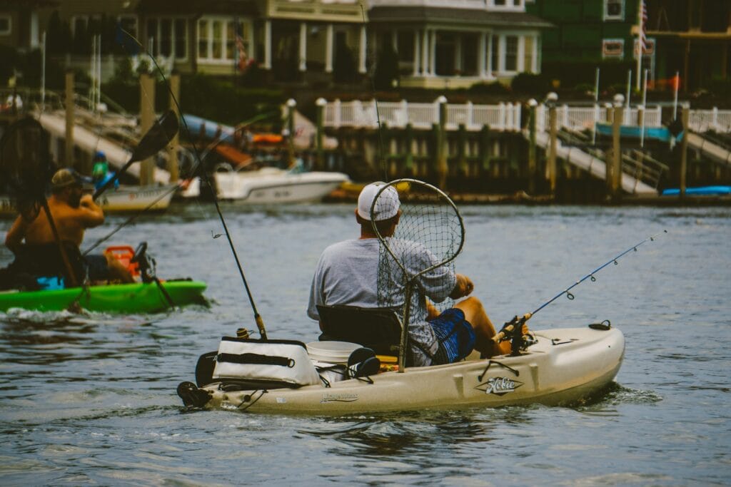 Two anglers paddle their kayaks in a calm waterway near a residential marina. The foreground features a man in a light-colored Hobie kayak, holding a fishing net while two fishing rods are secured on the kayak. In the background, another person in a green kayak is paddling nearby. The scene shows houses and boats docked along the waterfront, providing a serene setting for a day of fishing from a kayak. The overall atmosphere is peaceful and relaxed, with the kayakers focused on their activity.