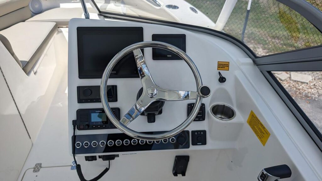 The helm of a Key West 239 DFS dual console boat, featuring a stainless steel steering wheel at the center. The dashboard is equipped with two large digital displays, controls for various boat functions, and a Garmin navigation system. Below the wheel are rows of switches for additional operations, while to the right is a cup holder for convenience. The layout is designed for easy access to all controls, providing both functionality and a sleek, modern look. The cockpit area in the background offers cushioned seating for passengers.