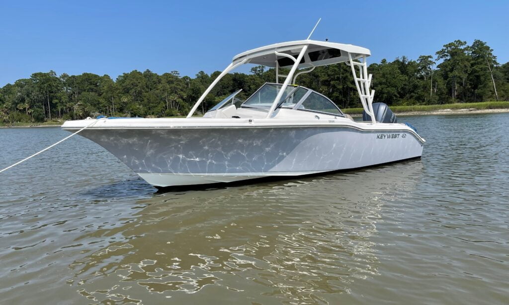 The image shows a Key West dual console boat floating on calm water, anchored near a shoreline. The boat has a white exterior with a hardtop roof and a sleek, modern design. It features a spacious bow and cockpit area, likely for seating and storage. The boat is equipped with an outboard engine at the back. In the background, there is a lush, tree-lined shoreline under a clear blue sky. The overall setting suggests a peaceful, scenic location, ideal for a day of boating.