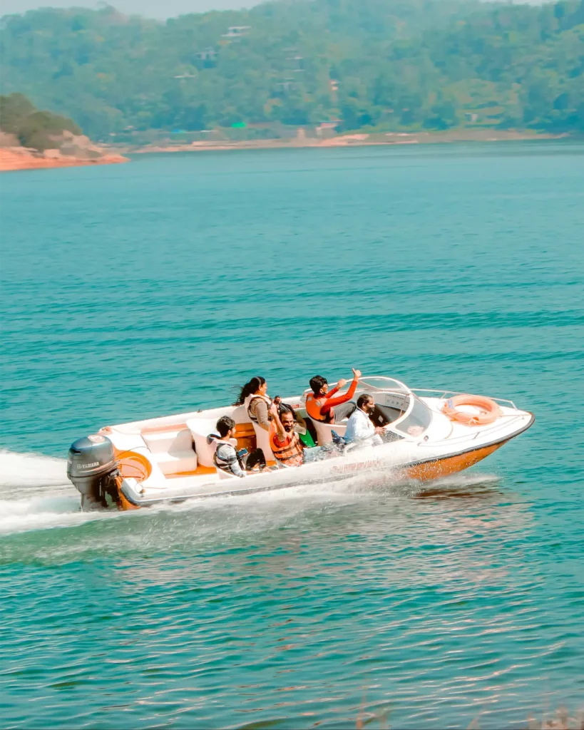 A group of people enjoying a boat ride on a clear, calm body of water. The boat is moving at a moderate speed, creating a small wake behind it. The passengers, wearing life jackets, are seated comfortably in the open boat, with one person raising their arm in excitement. The boat is powered by a Yamaha outboard motor and features bright orange and white accents. In the background, a lush green shoreline with hills and trees adds to the scenic view, creating a serene and joyful atmosphere on the water.