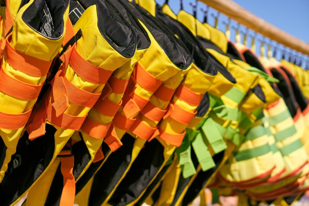 A row of brightly colored life jackets hanging on display, featuring a mix of yellow, black, and green fabrics with bold orange and green straps. The life jackets are neatly arranged on hangers, ready for use. The bright colors provide visibility and safety, making them ideal for boating or water activities. The image is set outdoors under a clear blue sky, adding to the sense of a sunny day spent near the water.