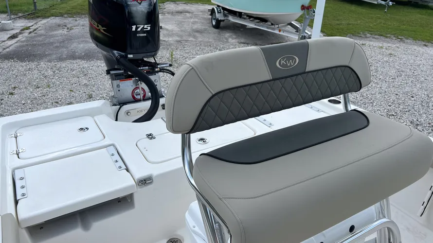 The leaning post seating on a Key West boat, featuring a cushioned backrest and seat in light gray with black accents. The backrest has a diamond-stitched pattern with the Key West (KW) logo embroidered in the center. The seat is positioned in front of the center console, providing a comfortable place for the captain to sit or lean while operating the boat. In the background, a Yamaha 175 outboard motor is mounted at the stern. The deck around the seating area is clean and organized, with storage hatches visible.