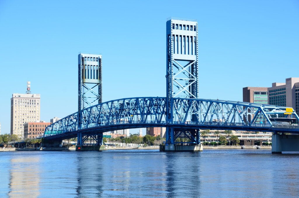 The iconic blue Main Street Bridge, also known as the John T. Alsop Jr. Bridge, spanning the St. Johns River in Jacksonville, Florida. The lift bridge is raised above the water, with its steel framework reflecting in the calm river below. Surrounding the bridge are several buildings, including historic and modern structures, with a clear blue sky overhead. The bridge serves as a prominent feature of the Jacksonville skyline and connects the downtown area to the Southbank.