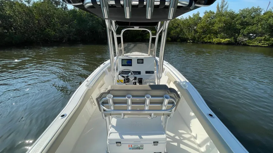 The center console area of a boat with a sleek, modern design. The helm features a stainless steel steering wheel, digital displays, and control switches for operating the boat. The T-top provides shade, and stainless steel rod holders are mounted on the back of the seat for easy access during fishing trips. The boat's deck is clean and spacious, offering plenty of room for movement. The calm water and shoreline with trees in the background create a peaceful atmosphere, ideal for cruising or fishing.