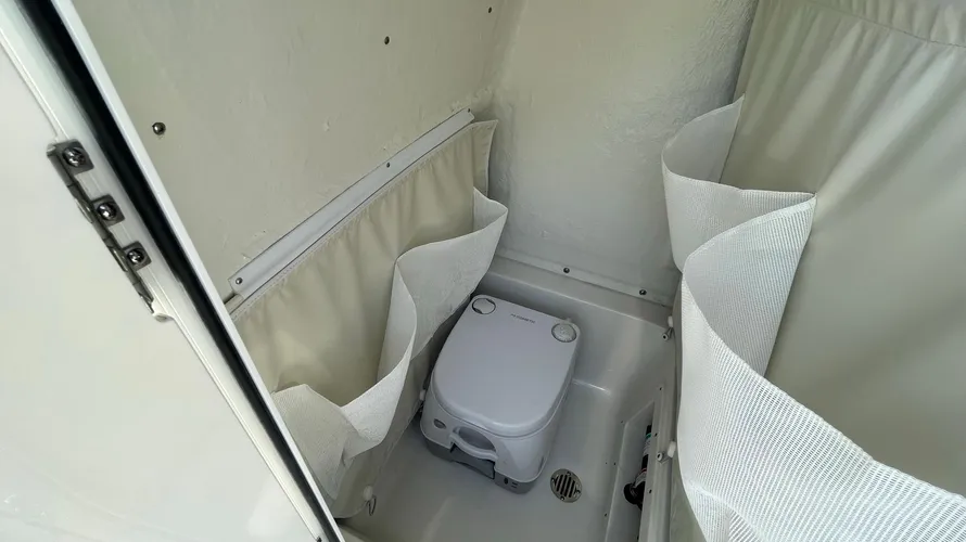 A compact marine head compartment on a boat, featuring a portable toilet with simple controls. The walls of the compartment are equipped with storage pouches for holding small items. The space is designed to be functional, with a small drain on the floor for easy cleaning. The walls are light-colored, and the compartment is enclosed, providing privacy for passengers. The setup is practical for boaters needing a restroom option while out on the water.