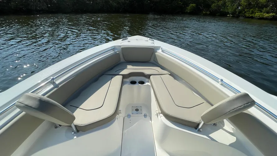 The bow seating area of a boat, featuring cushioned seats in light beige with backrests on both sides. The seating is arranged in a V-shape, providing comfortable space for passengers to relax at the front of the boat. Two cup holders are located in the center for convenience. Stainless steel grab rails run along the sides for added safety. The boat is floating in calm water, with trees visible along the shoreline in the background, creating a serene and relaxing setting for cruising or anchoring.