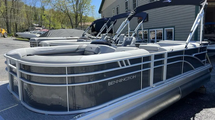 The image shows a Bennington pontoon boat with a sleek gray and black exterior. It features plush seating, a spacious deck layout, and a dual canopy for shade. The boat is docked outside on a trailer in front of a building, with trees and another covered boat in the background. The Bennington brand name is prominently displayed on the side of the boat.






