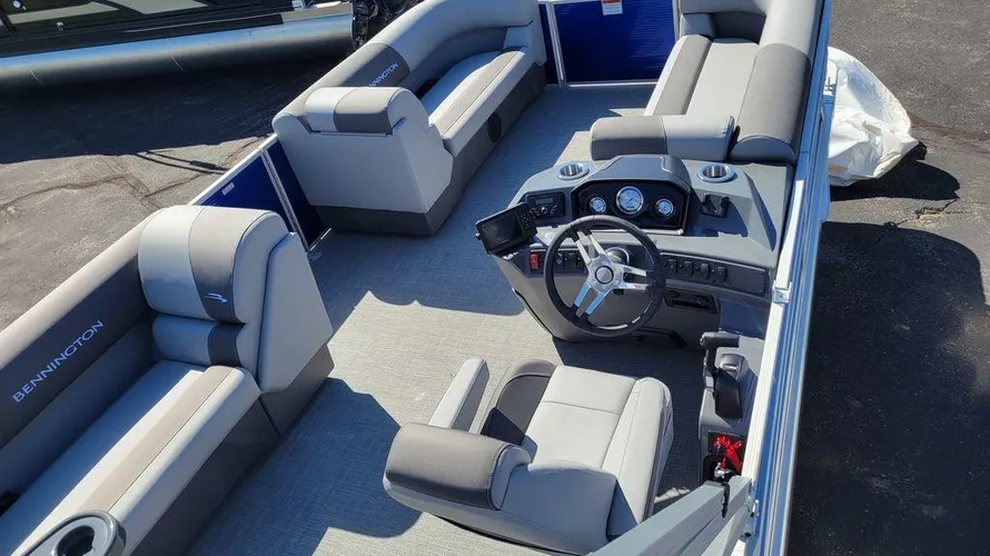 The interior of a Bennington pontoon boat, featuring spacious seating areas with cushioned benches in light gray and darker accents. The captain's helm includes a sleek steering wheel, control panel, and navigation screen, all designed for ease of use. The boat's layout is open and inviting, with ample seating for passengers along both sides and the rear. Cup holders are integrated into the seating areas for convenience. The flooring is a light-colored, durable material, contributing to the comfortable and relaxed environment ideal for cruising or entertaining on the water.