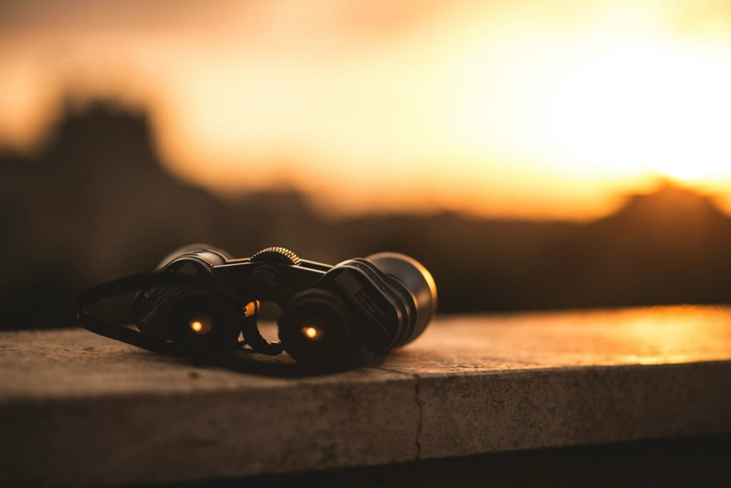 A pair of binoculars resting on a stone surface, captured during a golden sunset. The warm light from the setting sun casts a soft glow on the binoculars, highlighting their sleek design. The background is blurred, with silhouettes of distant trees and buildings blending into the horizon, enhancing the serene and peaceful mood of the scene. The image evokes a sense of exploration or quiet observation, with the binoculars symbolizing a moment of reflection or anticipation.