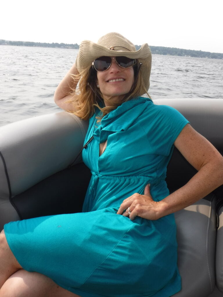 A woman relaxing on a boat, wearing a turquoise dress, a cowboy-style hat, and sunglasses. She is seated comfortably on the cushioned seating of the boat, smiling and enjoying the breeze. In the background, a calm body of water and a distant shoreline are visible, creating a peaceful and serene atmosphere. The woman’s casual, relaxed posture and smile reflect a laid-back day on the water.