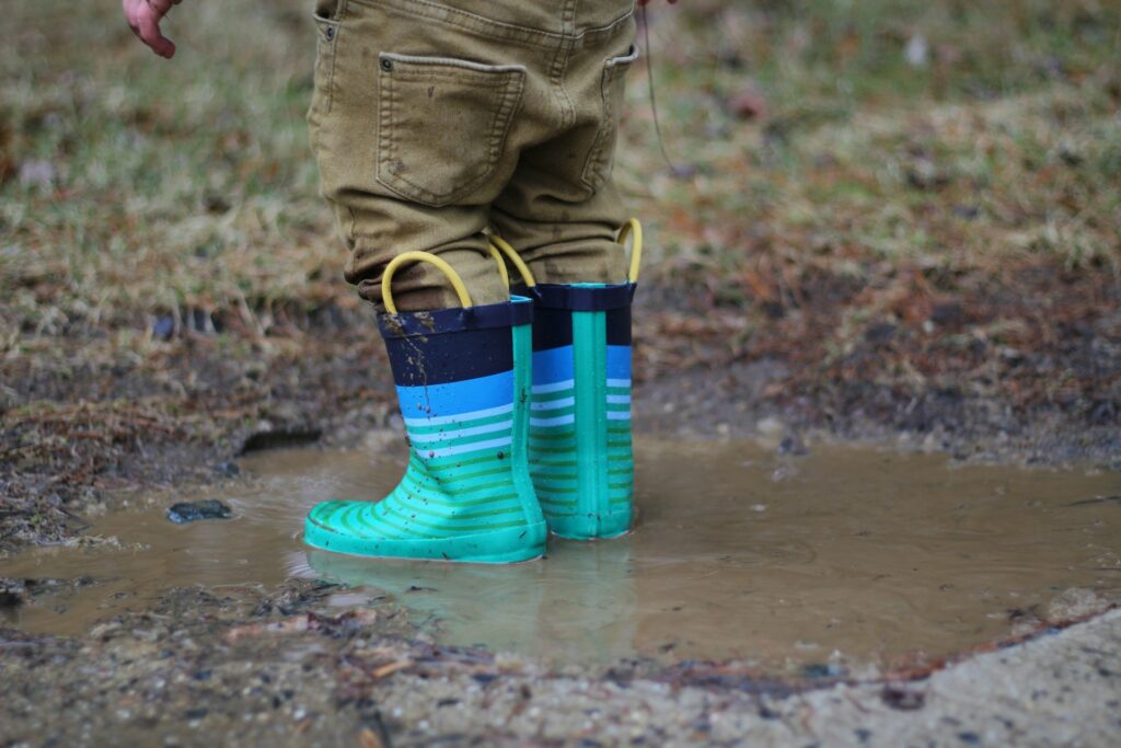 A child standing in a muddy puddle, wearing bright blue and green striped rain boots. The child is dressed in tan pants, and only their lower legs are visible. The boots are splashing in the water, which is surrounded by wet grass and dirt. The scene captures a playful, carefree moment outdoors, where the child is enjoying the simple joy of jumping in puddles after a rain. The background is slightly blurred, focusing on the boots and the muddy ground.