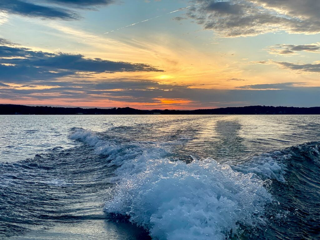 A scenic view from the back of a boat, capturing the wake left behind as the boat moves across a calm lake at sunset. The sky is painted with vibrant hues of orange, pink, and blue as the sun sets on the horizon. Dark silhouettes of distant landmasses are visible against the colorful sky, creating a serene and peaceful atmosphere on the water.