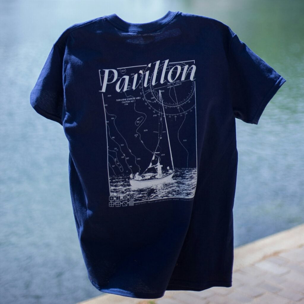 A navy blue t-shirt hanging against a backdrop of water, featuring a white graphic design on the back. The design includes nautical chart details, with contour lines and a sailboat at sea, and the word "Pavillon" prominently displayed at the top. The image gives off a maritime theme, evoking a sense of adventure and the open ocean. The shirt is displayed in a way that highlights the intricate and detailed design, with the water in the background reinforcing the nautical vibe.