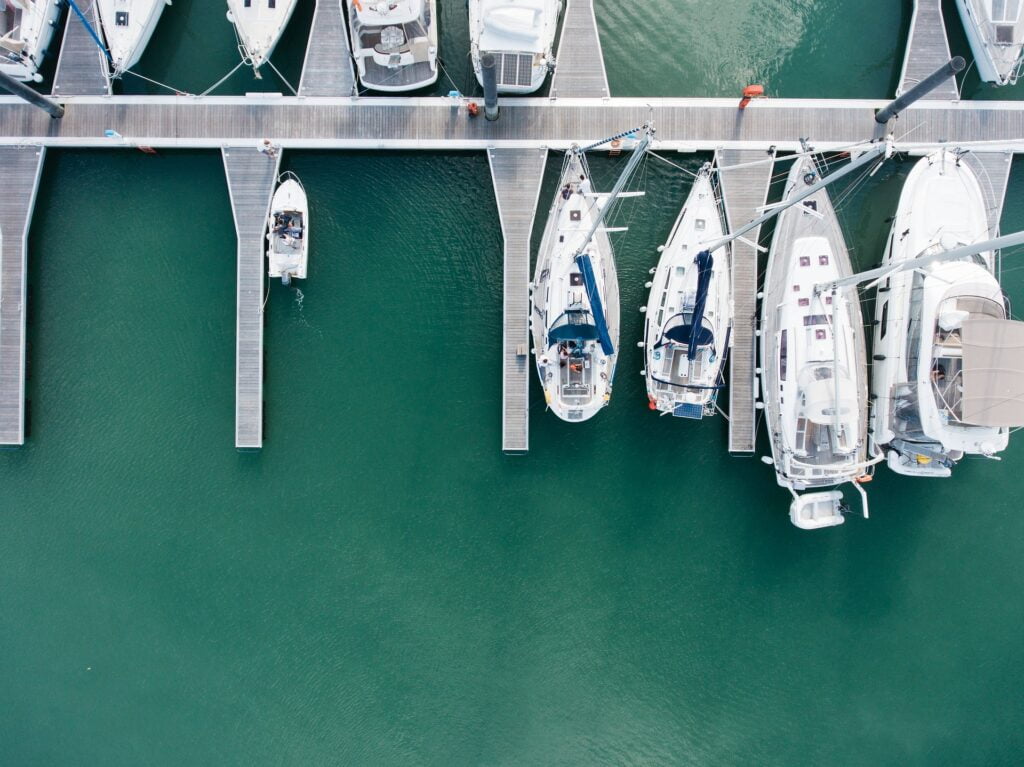 An aerial view of a marina showing several sailboats and yachts docked along wooden piers in calm, green water. The boats are neatly lined up in individual berths, with walkways extending out to each vessel. A small motorboat can be seen maneuvering away from the dock. The clear, organized layout of the marina highlights the orderly arrangement of boats, and the serene water gives the scene a peaceful and quiet atmosphere, ideal for a day at the harbor.