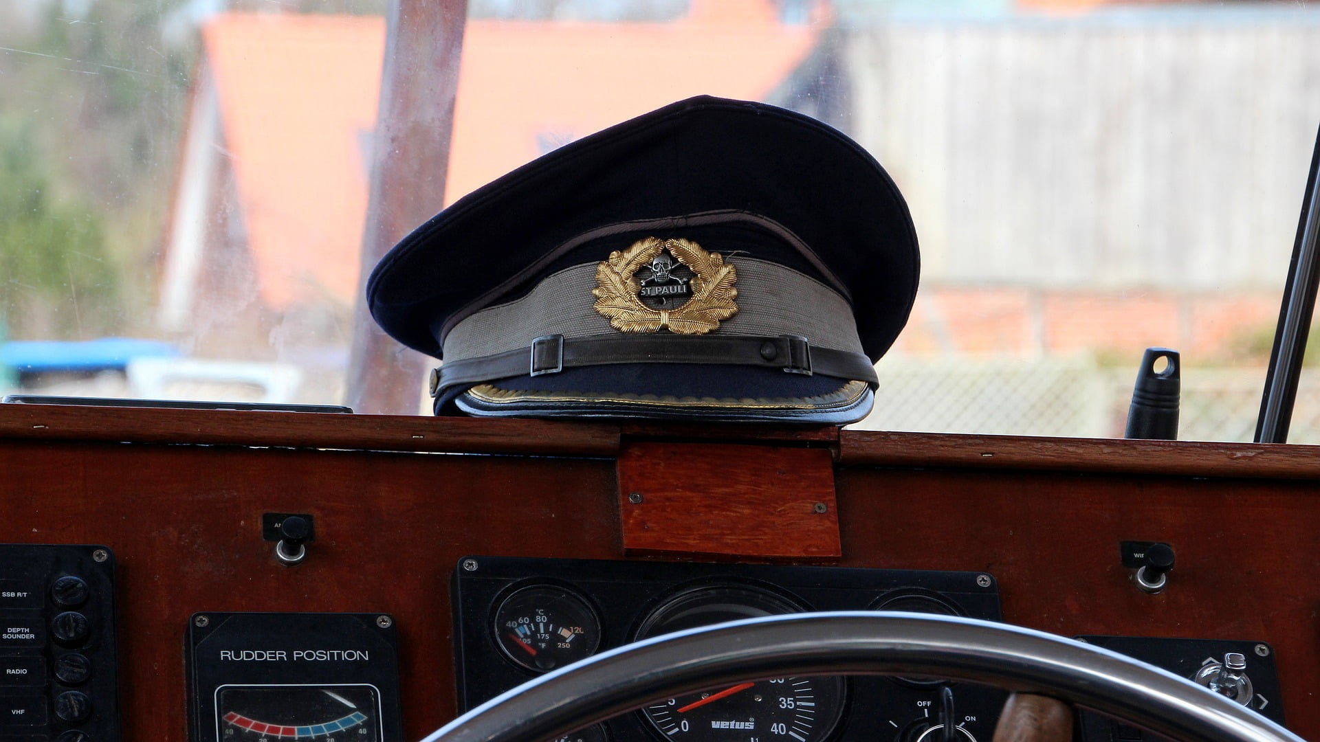 A captain's hat resting on the dashboard of a boat, positioned above the instrument panel. The hat is navy blue with a silver band and features a decorative gold emblem in the center, marked with the words "St. Pauli." Below the hat, the boat's dashboard includes various gauges, including one labeled "Rudder Position." The setting suggests a classic or vintage boat, with wooden details adding to the traditional maritime atmosphere. The blurred background through the window hints at a dock or harbor area.