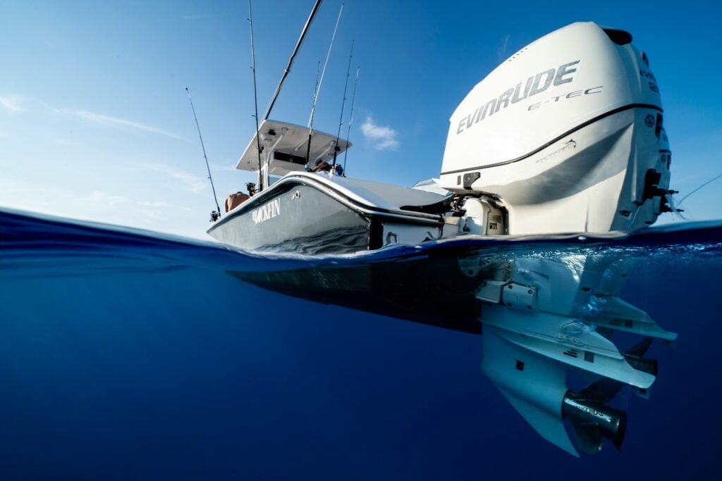 A dramatic split-level shot of a fishing boat floating on the ocean, partially submerged. The underwater portion reveals the boat's outboard Evinrude E-TEC engine, while the top half shows the boat's deck and fishing rods. The boat is named 'OAKEN,' and the image captures the clear blue water and a bright blue sky above.