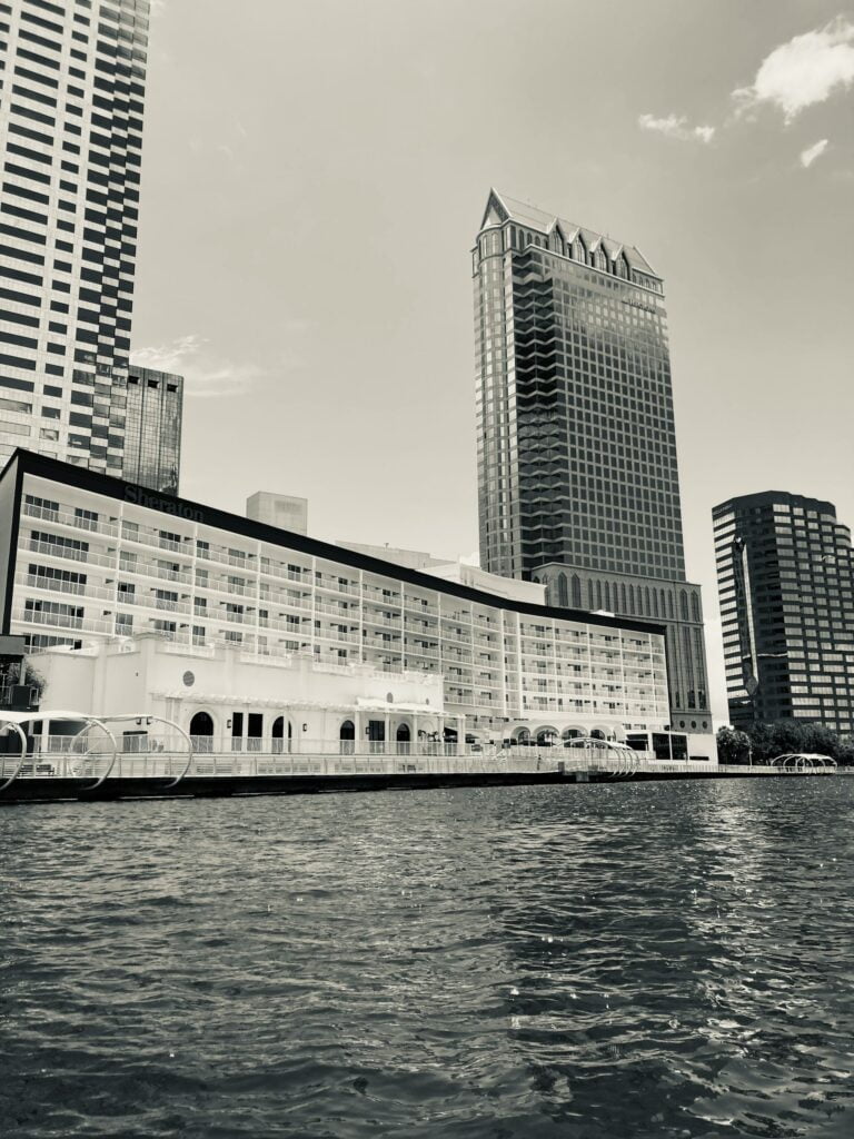 A black and white image of a waterfront cityscape featuring tall buildings, including a prominent high-rise with a distinctive pointed roof and a Sheraton hotel along the water. The view is from the water, with gentle ripples in the foreground, and a mix of modern and classic architectural styles in the background. The sky is mostly clear with a few clouds visible.