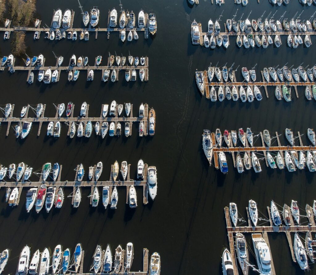 An aerial view of a busy marina with numerous boats docked along parallel piers. The boats are neatly arranged in rows, with a variety of vessels, including sailboats and motorboats, occupying the slips. The water is dark, and the piers extend out into the marina from the shore, providing a clear view of the boats and their arrangement.