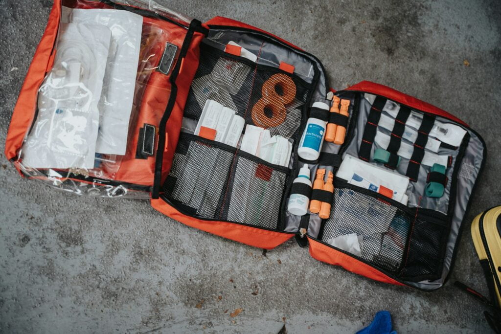 An open, well-organized first aid kit displayed on the ground. The kit is housed in a bright red case with several compartments, each containing essential medical supplies. Visible items include bandages, gauze, antiseptic bottles, and medical tape. The compartments are made of mesh, allowing for easy visibility and access to the items inside. The kit appears comprehensive and is designed for emergency situations, ideal for outdoor activities or travel. The overall appearance suggests preparedness for medical emergencies.