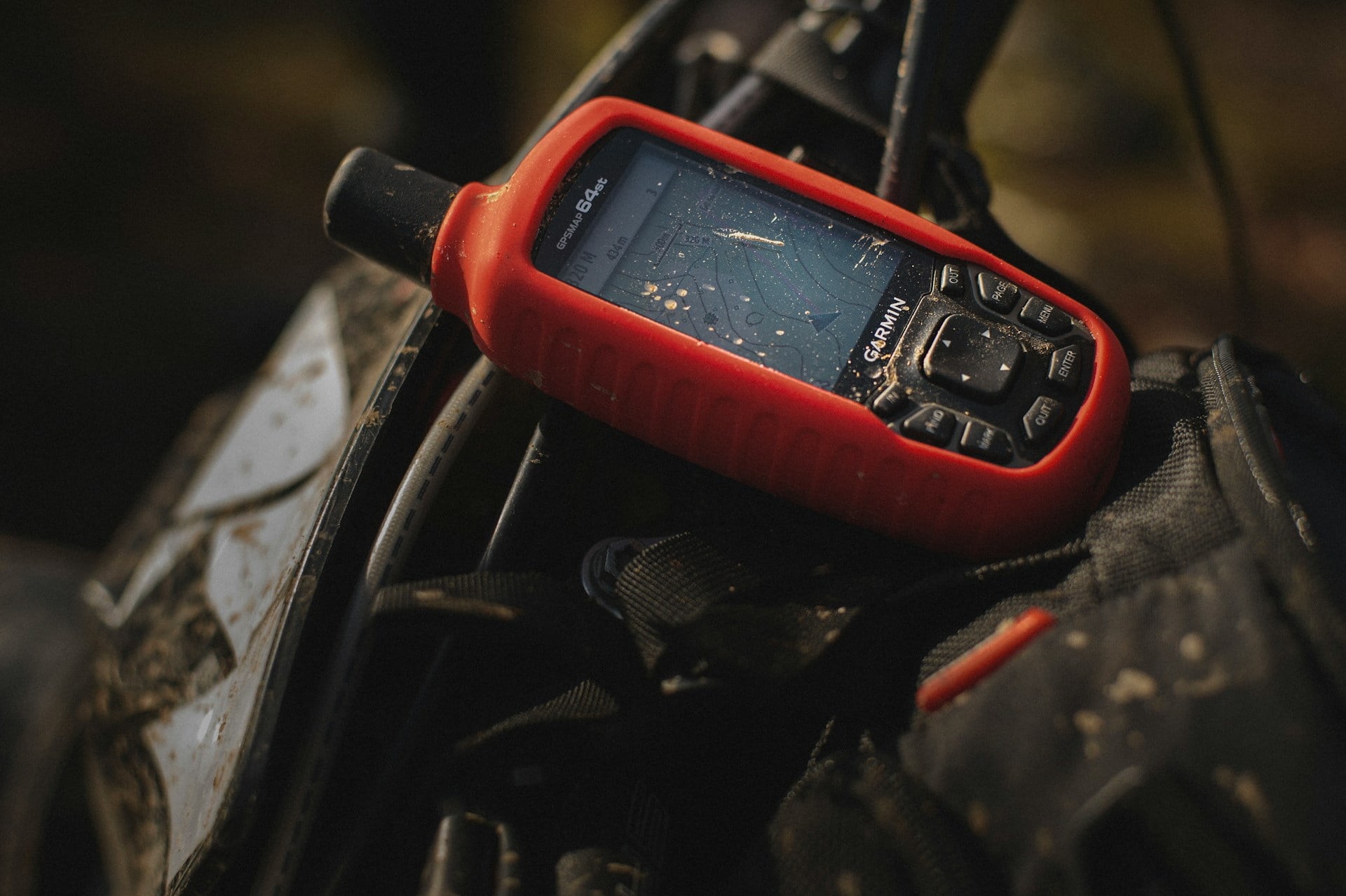 A close-up of a Garmin GPS device in a rugged red case, resting on a backpack outdoors. The screen shows topographical map details, indicating its use for navigation. The device appears slightly dusty, suggesting recent use in an outdoor or off-road setting. The surrounding gear, including straps and buckles, reflects an adventure-ready environment, ideal for hiking, camping, or exploring in remote locations. The scene conveys a sense of preparedness and outdoor exploration.