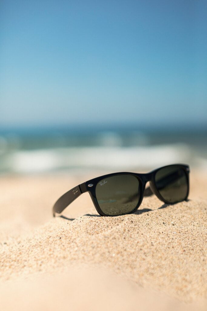 A pair of black Ray-Ban sunglasses resting on golden sand at the beach. The iconic sunglasses are positioned at an angle, with the Ray-Ban logo visible on the side. In the background, the ocean is slightly out of focus, with gentle waves and a clear blue sky adding to the peaceful beach setting. The image evokes a sense of relaxation and enjoying a sunny day by the water.