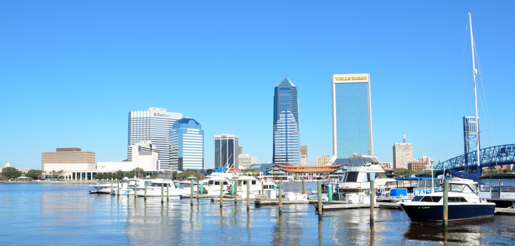 A bright, clear day view of the Jacksonville, Florida, waterfront. In the foreground, several boats are docked at a marina, with calm waters reflecting their hulls. The skyline of downtown Jacksonville dominates the background, showcasing modern skyscrapers like the Wells Fargo building and the Bank of America tower. The iconic blue Main Street Bridge (John T. Alsop Jr. Bridge) is visible on the right side, spanning the St. Johns River. The cityscape contrasts with the tranquil marina, creating a vibrant urban-meets-waterfront scene.