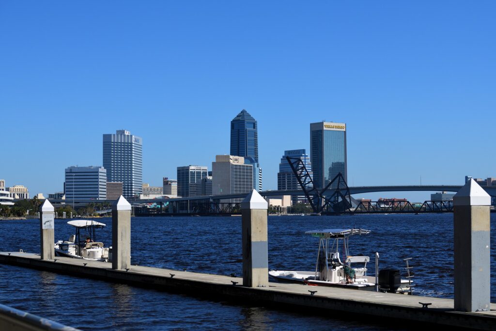 A scenic view of the Jacksonville skyline from the waterfront, featuring modern skyscrapers, including the Wells Fargo building and other notable buildings. In the foreground, boats are docked along a pier with white posts. The blue sky and calm waters of the river provide a serene backdrop, with a bridge visible in the distance.