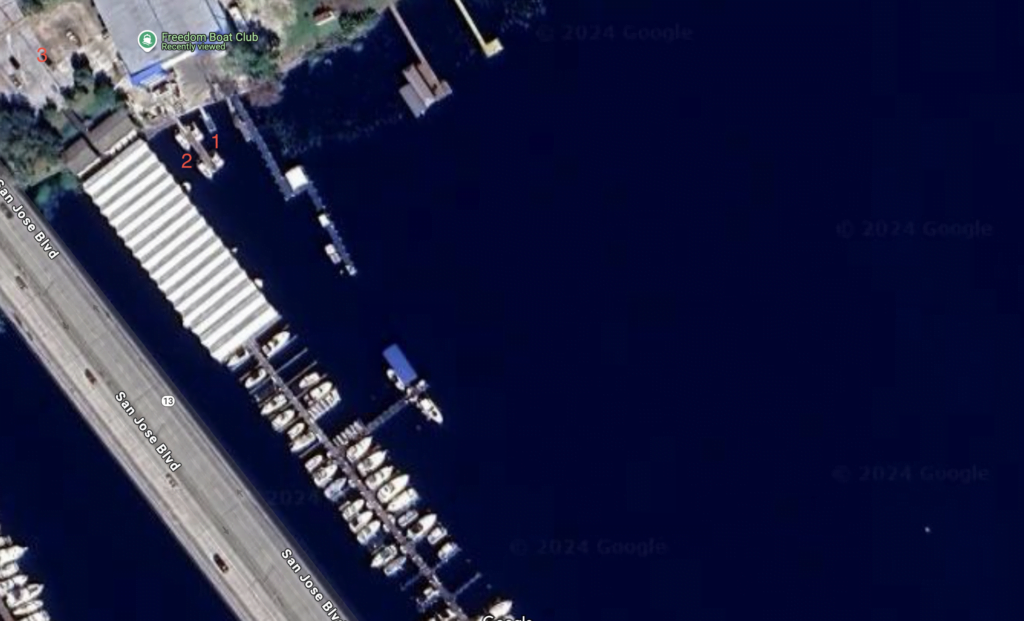 An aerial satellite view of a marina located along San Jose Boulevard. The image shows boats docked along multiple piers and covered boat storage facilities on the left. The marina sits next to a highway, labeled San Jose Boulevard, which runs along the left side of the image. Several boats are moored at the docks, and key areas are marked with numbers for reference, likely indicating specific facilities or areas of interest. The deep blue water contrasts with the lighter structures of the docks and boats.