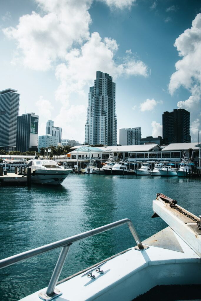 A view from a boat docked in a marina, overlooking a skyline of modern high-rise buildings. Several yachts and boats are moored at the docks in the foreground, while the towering skyscrapers rise into the sky with a mix of white clouds. The water is calm, reflecting the surrounding structures, and the marina appears to be part of a busy urban waterfront.