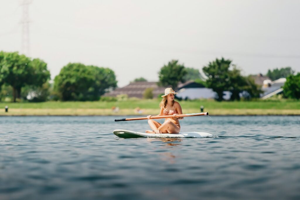 A woman sitting on a paddleboard in calm water, holding a paddle across her lap. She is wearing a sunhat and a swimsuit, enjoying the peaceful scenery. The background shows a grassy shoreline with trees and distant houses under a clear sky.