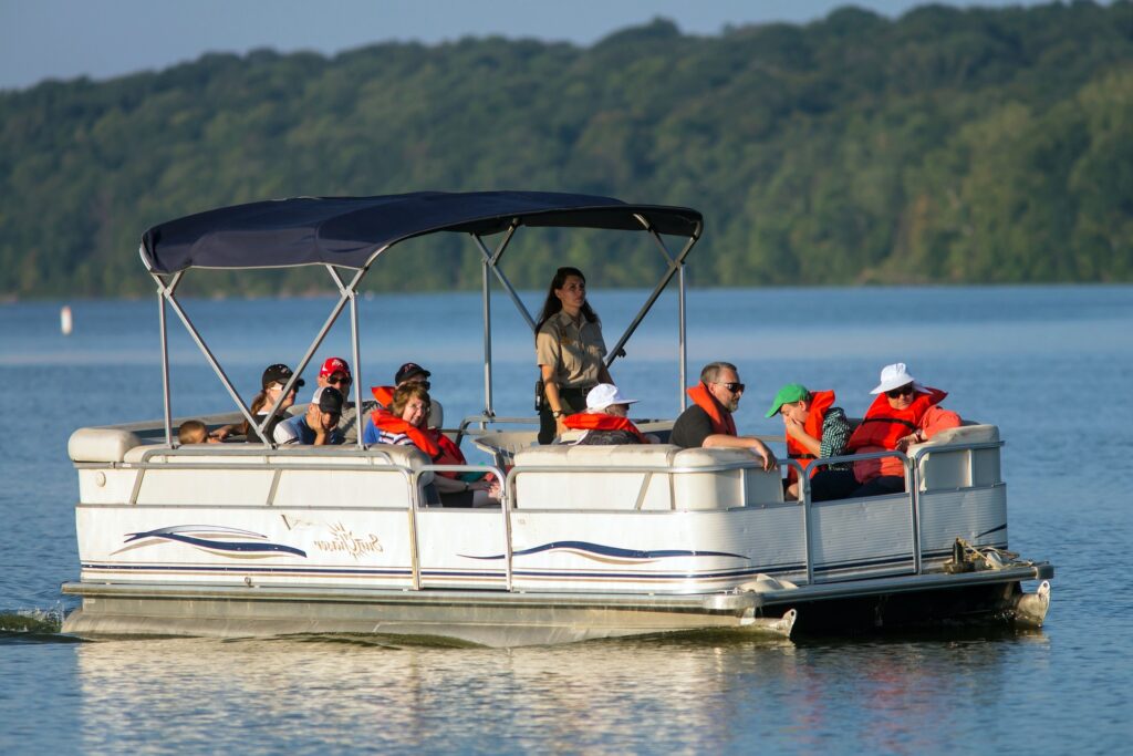 A group of people enjoy a relaxing ride on a pontoon boat on a calm lake. Everyone on board is wearing life jackets, and the boat is shaded by a canopy. The boat operator is standing at the helm. In the background, a wooded shoreline is visible under a soft, clear sky. The atmosphere appears peaceful as the boat glides over the water.