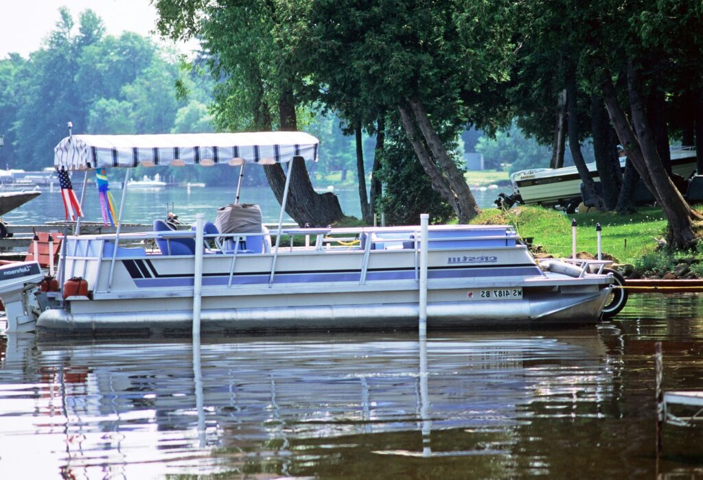 A pontoon boat is docked near a shoreline, surrounded by calm water and greenery. The boat features a blue and white striped canopy for shade and comfortable seating. In the background, tall trees and other boats create a serene lakeside scene. The boat has an American flag at the stern, gently waving in the breeze. The atmosphere is peaceful, with reflections of the boat and trees on the water.