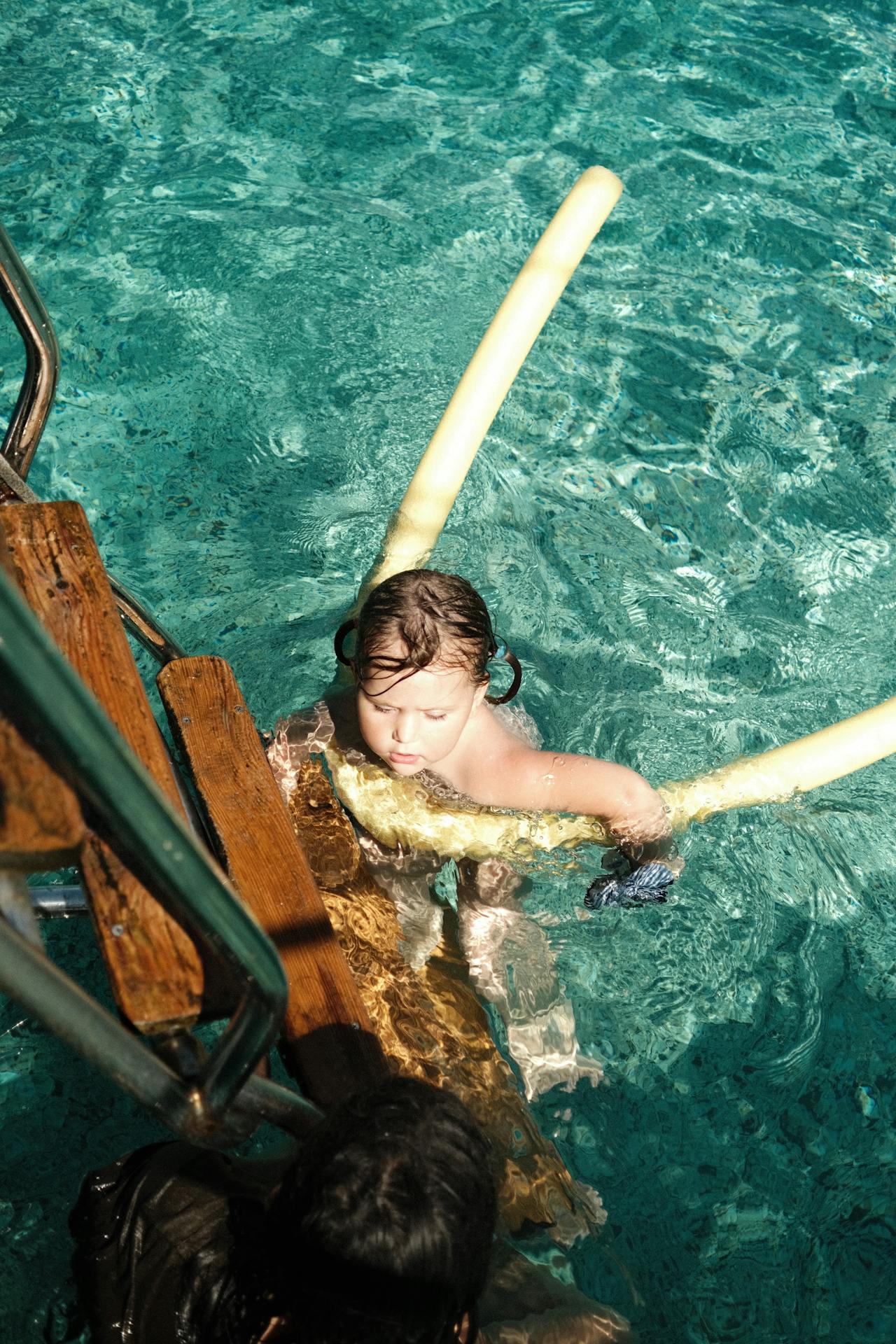 A young child swimming in a pool, holding onto a yellow pool noodle for support. The child is near a wooden pool ladder, using it to steady themselves while in the water. The clear blue water of the pool shimmers in the sunlight, creating a refreshing and playful scene. The child appears focused and is enjoying a swim, with a darker-haired person visible at the bottom of the image, possibly supervising or helping. The scene evokes a sense of fun and safety during a day of swimming.