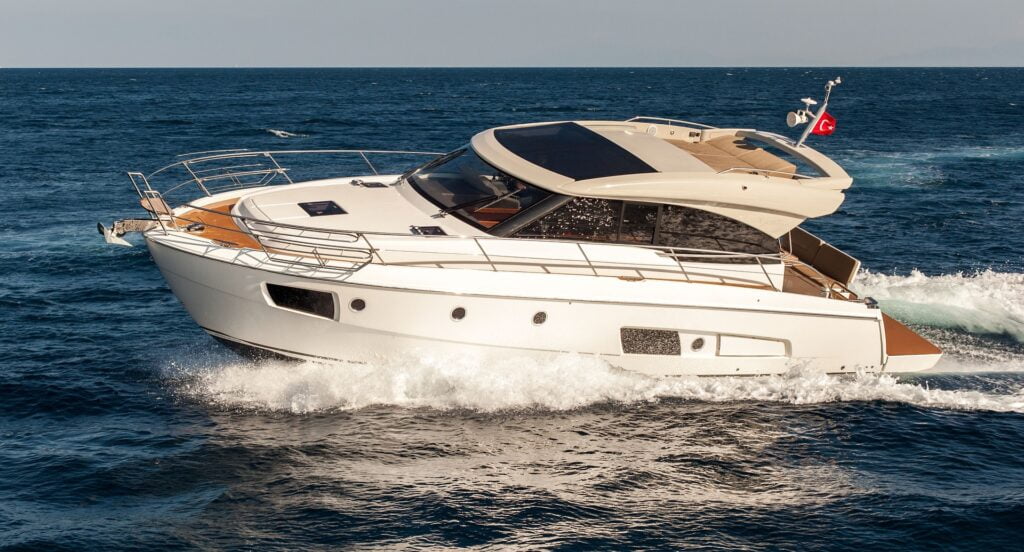 A sleek white motor yacht cruising through open waters, with waves splashing against its hull. The yacht features a modern design with a streamlined shape, large windows, and an upper deck covered by a sunroof. A Turkish flag is seen flying from the rear of the vessel. The boat appears to be moving at a fast speed, with a clear blue sky and expansive ocean surrounding it. The image captures the yacht in motion, creating a sense of luxury and adventure on the water.