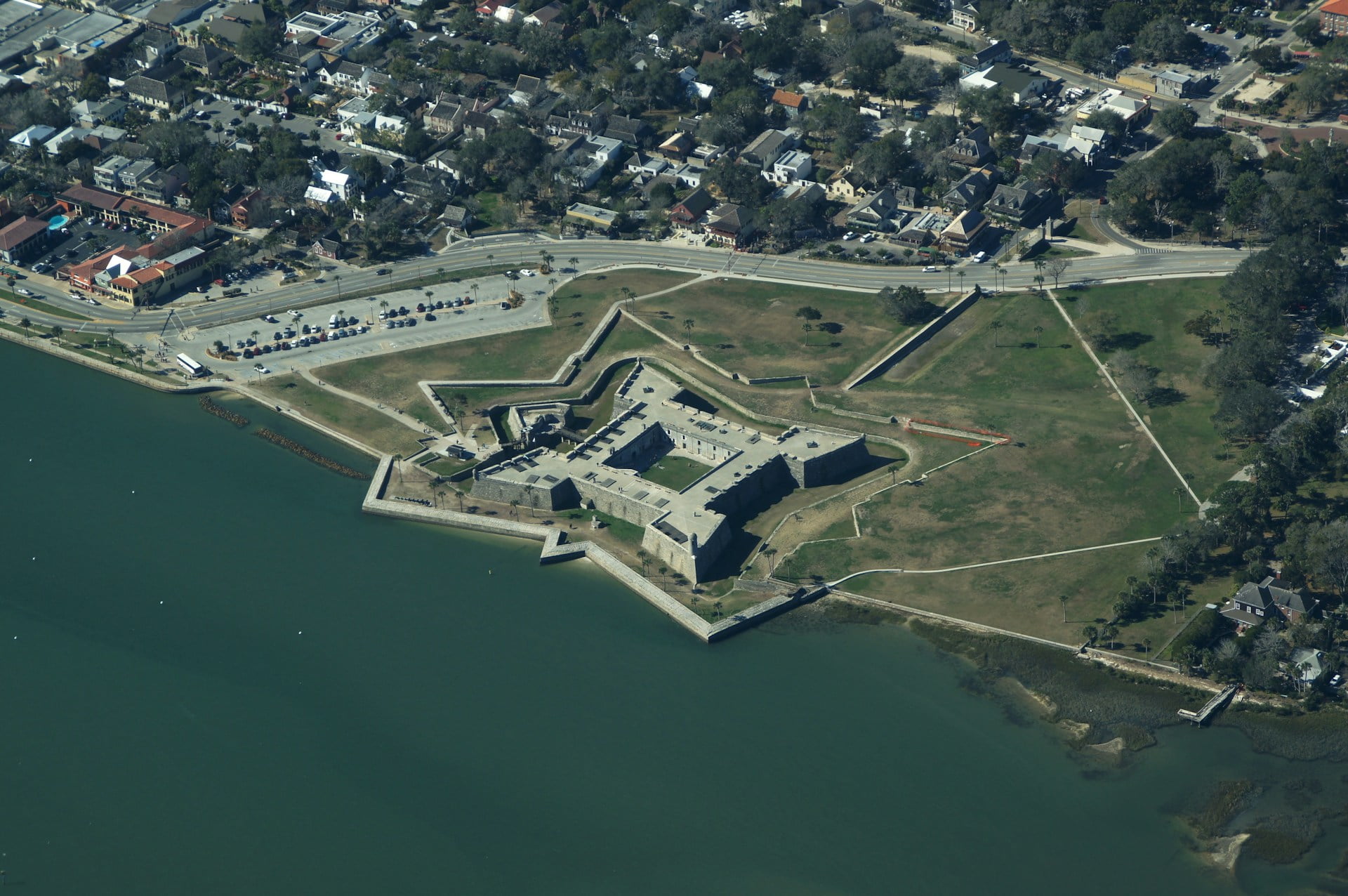 An aerial view of Castillo de San Marcos, the historic fort in St. Augustine, Florida. The star-shaped structure, built of coquina stone, stands on the waterfront of Matanzas Bay, surrounded by lush green lawns and a stone seawall. The fort's well-preserved walls and battlements form a geometric pattern, typical of European fortress designs of the colonial era. Nearby, you can see the town’s layout with buildings and streets surrounding the historical site. The bay waters are calm, reflecting the fort's proximity to the coastline. This iconic fort is a significant symbol of St. Augustine's Spanish colonial past.