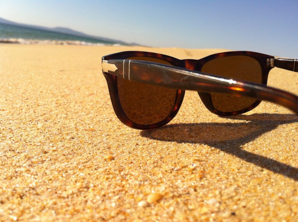 A pair of tortoiseshell sunglasses resting on a golden sandy beach under bright sunlight. The sand is fine, with small grains visible in the foreground, and the clear blue sky stretches into the distance. In the background, the ocean waves gently lap the shore, with distant hills faintly visible along the horizon. The scene evokes a relaxed, sunny day at the beach, with the sunglasses symbolizing a laid-back, carefree atmosphere.