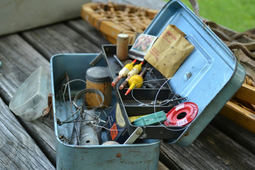 An open, vintage fishing tackle box resting on a wooden surface, filled with a variety of fishing gear. The box contains fishing lines, bobbers, hooks, a spool of fishing thread, and small containers holding other accessories. The weathered blue metal box has compartments to keep the gear organized. Nearby, a wicker basket can be seen, adding to the rustic, outdoor setting. The scene evokes a nostalgic feel, suggesting preparation for a day of fishing by the lake or river.