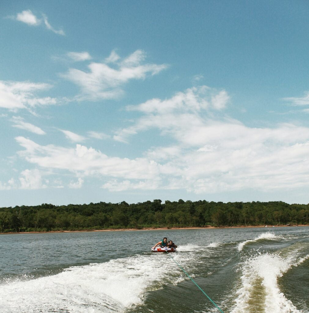 Two people riding on an inflatable tube being pulled by a boat across a lake. The tube skims over the water's surface, creating a wake behind it as the boat moves forward. The riders are holding on and enjoying the thrill of tubing. In the background, a tree-lined shoreline under a blue sky with scattered clouds provides a scenic backdrop. The image captures the excitement and fun of watersports on a calm day, with the open water and natural surroundings adding to the adventurous atmosphere.