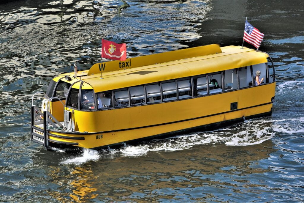 A bright yellow water taxi moves along a river, featuring an American flag and a Marine Corps flag on its roof. The boat is designed like a small bus, with large windows and seating visible inside. One passenger can be seen sitting near the back, looking at their phone. The water taxi is cutting through the calm, reflective waters as it travels.