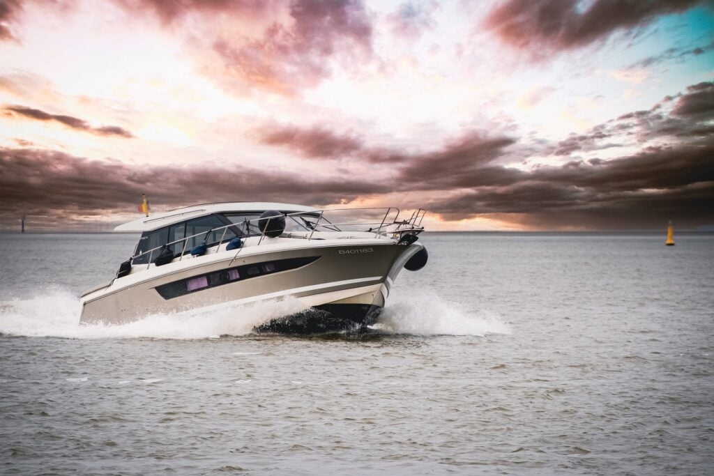 A sleek motor yacht cruising through calm waters under a dramatic sunset sky. The boat's bow cuts through the water, creating white waves, while the sky is filled with dark, moody clouds tinged with pink, purple, and orange hues from the setting sun. The modern design of the yacht contrasts with the natural beauty of the scene, emphasizing a sense of adventure and luxury on the open water.