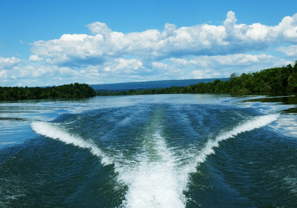 The image shows a calm, expansive body of water with a boat's wake cutting through the center. The water is smooth with minimal ripples, and the wake creates two distinct trails curving away from the boat. The scenery is framed by lush green trees on both sides, leading into the distance where a range of low-lying mountains meets a blue sky filled with fluffy white clouds. The image captures the tranquility of a boating day in a peaceful, natural environment.