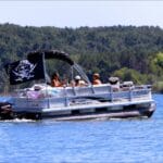 The image shows a group of people relaxing on a pontoon boat in calm waters. The boat is a Sun Tracker model, powered by a Mercury outboard motor, and is adorned with a pirate flag featuring a skull and crossed swords. The group appears to be enjoying a leisurely outing on a sunny day, surrounded by a backdrop of green trees lining the distant shore. The boat has comfortable seating, with some inflatable tubes visible on board, suggesting they may be preparing for or have finished enjoying water activities.