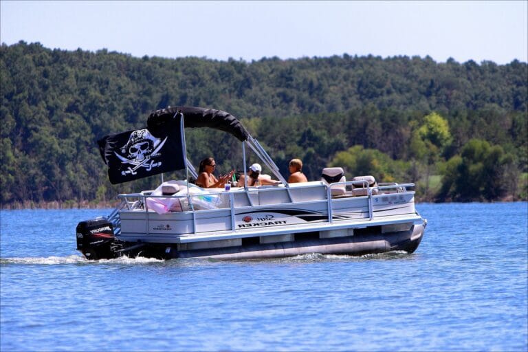 The image shows a group of people relaxing on a pontoon boat in calm waters. The boat is a Sun Tracker model, powered by a Mercury outboard motor, and is adorned with a pirate flag featuring a skull and crossed swords. The group appears to be enjoying a leisurely outing on a sunny day, surrounded by a backdrop of green trees lining the distant shore. The boat has comfortable seating, with some inflatable tubes visible on board, suggesting they may be preparing for or have finished enjoying water activities.