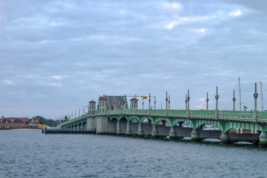 Image of the Bridge of Lions in St. Augustine, Florida, stretching across the Matanzas River under a cloudy sky. The green bridge features its signature towers and ornate street lamps, with part of the drawbridge raised. Sailboats are visible in the background, and the waterfront buildings line the horizon.