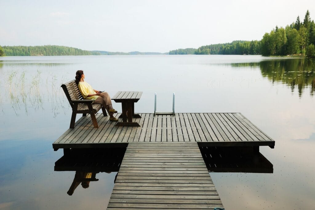 The image shows a peaceful scene of a person sitting on a wooden bench at the edge of a dock, gazing out over a calm, reflective lake. The dock extends into the water, with a small table next to the bench. Surrounding the lake is a dense forest of green trees, creating a serene and tranquil environment. The person appears to be relaxing and enjoying the quiet, natural beauty of the setting, with still water mirroring the sky and trees. The overall atmosphere is one of solitude and peacefulness in nature.