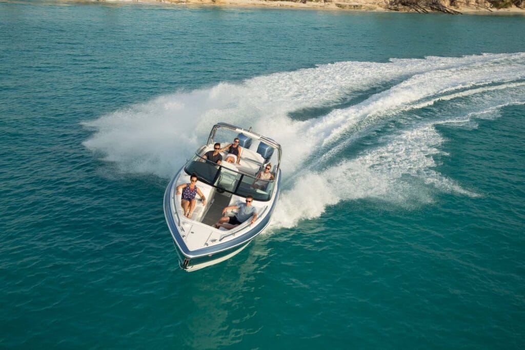 A speedboat with multiple passengers glides through clear turquoise waters, leaving a wide trail of white wake behind. The passengers are comfortably seated and enjoying the ride under sunny skies near a sandy shoreline.