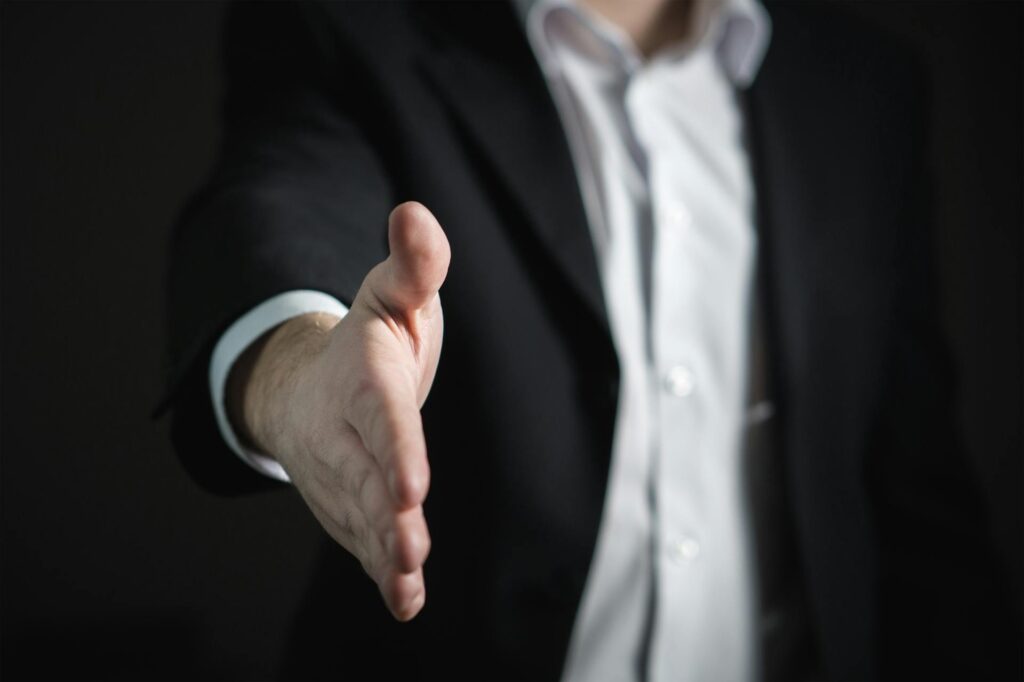 Close-up of a person wearing a dark suit and white shirt, extending their hand forward as if offering a handshake. The background is dark, emphasizing the outstretched hand in the foreground, symbolizing an offer, agreement, or business deal.